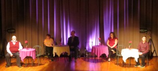 (Image: Cast Members Seated at Tables with Coloured Lighting on
  the Back Curtain)